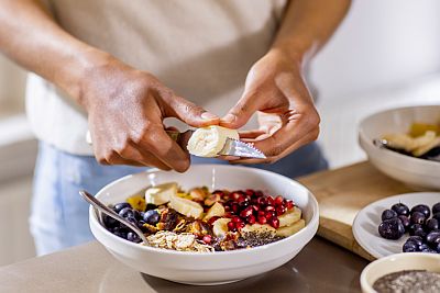 cutting banana over a breakfast bowl with yogurt, berries, chia seeds, and nuts